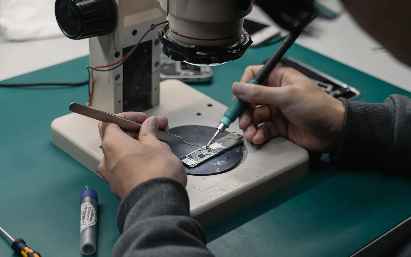 Technicien penche sur un microscope stereo, fer a souder et pince sur une carte mere de smartphone