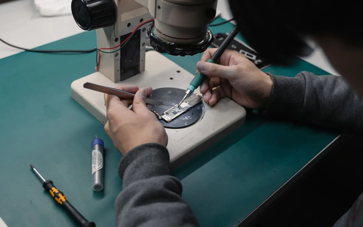 Mains tenant une pince et un fer à souder fin sur une carte mère, sous l'objectif du microscope
